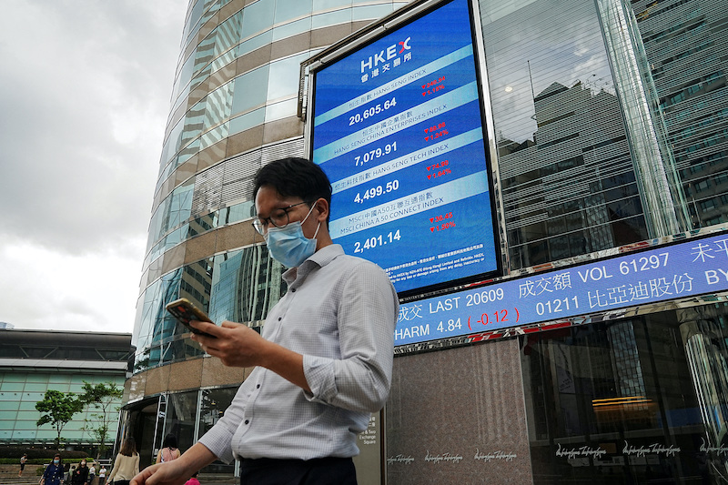 A man looks at his phone as people walk past a screen with the Hang Seng stock index outside Hong Kong Exchanges