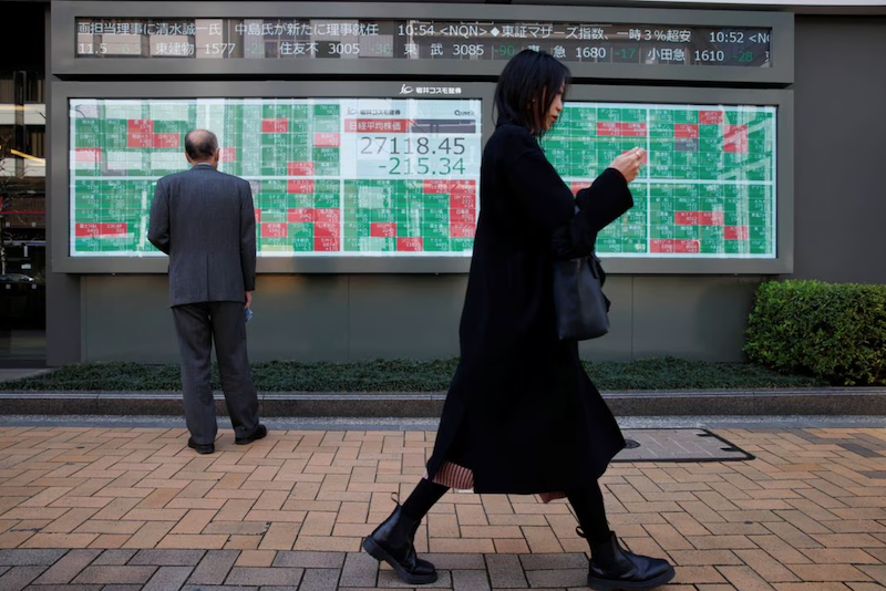 A woman walks past a man examining an electronic board showing Japan's Nikkei average and stock quotations outside a brokerage, in Tokyo, Japan, March 20, 2023. REUTERS/Androniki Christodoulou/File Photo